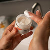 A close-up of a person's hands holding an open jar of white cream and applying it with a spatula. The jar has a white label with orange and black text, and the brand name 'Plantd'