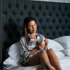 A woman sits on a bed in a bedroom, wearing a light gray satin shirt and holding a small cup. The bed has a large dark gray tufted headboard, white bedding, a wooden nightstand with a lamp and books, and birch tree patterned wallpaper.