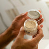 A person's hands holding an open jar of a light beige cosmetic or skincare product, with the lid resting beside it, against a softly blurred bathroom background.