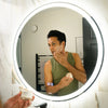 A man applies skincare cream to his face while looking into a large, illuminated round bathroom mirror. He is wearing an olive green tank top and holding a small jar of the cream in the foreground.
