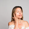 A woman with closed eyes applies a white cream from a small white jar to her cheek, wearing a white off-the-shoulder lace top against a plain light gray background.