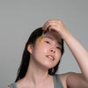 A woman with freckles applies a golden-yellow serum dropper to her forehead against a solid gray background.