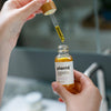 A close-up of a person holding a small glass dropper bottle labeled "plantd" while dropping golden-yellow serum onto a surface in a bathroom setting.