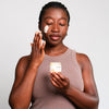 A woman applies a cream to her face holding a small jar of the product in her other hand, set against a plain light gray background.