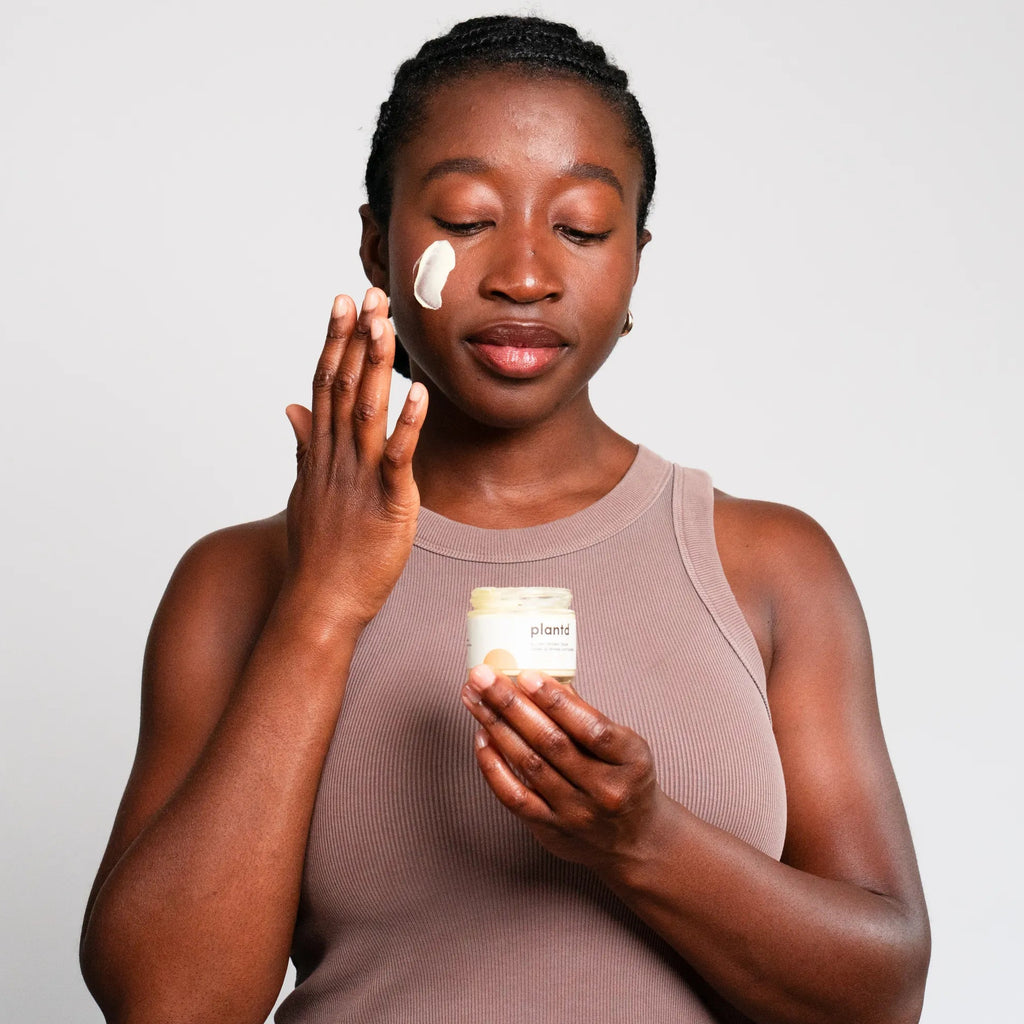 A woman applies a cream to her face holding a small jar of the product in her other hand, set against a plain light gray background.