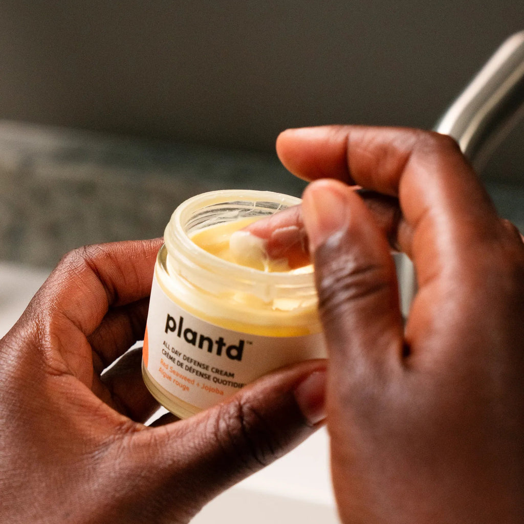 A close-up of a person's hands holding an open jar of Plantd hair serum and applying a dollop of the yellow cream with a finger.