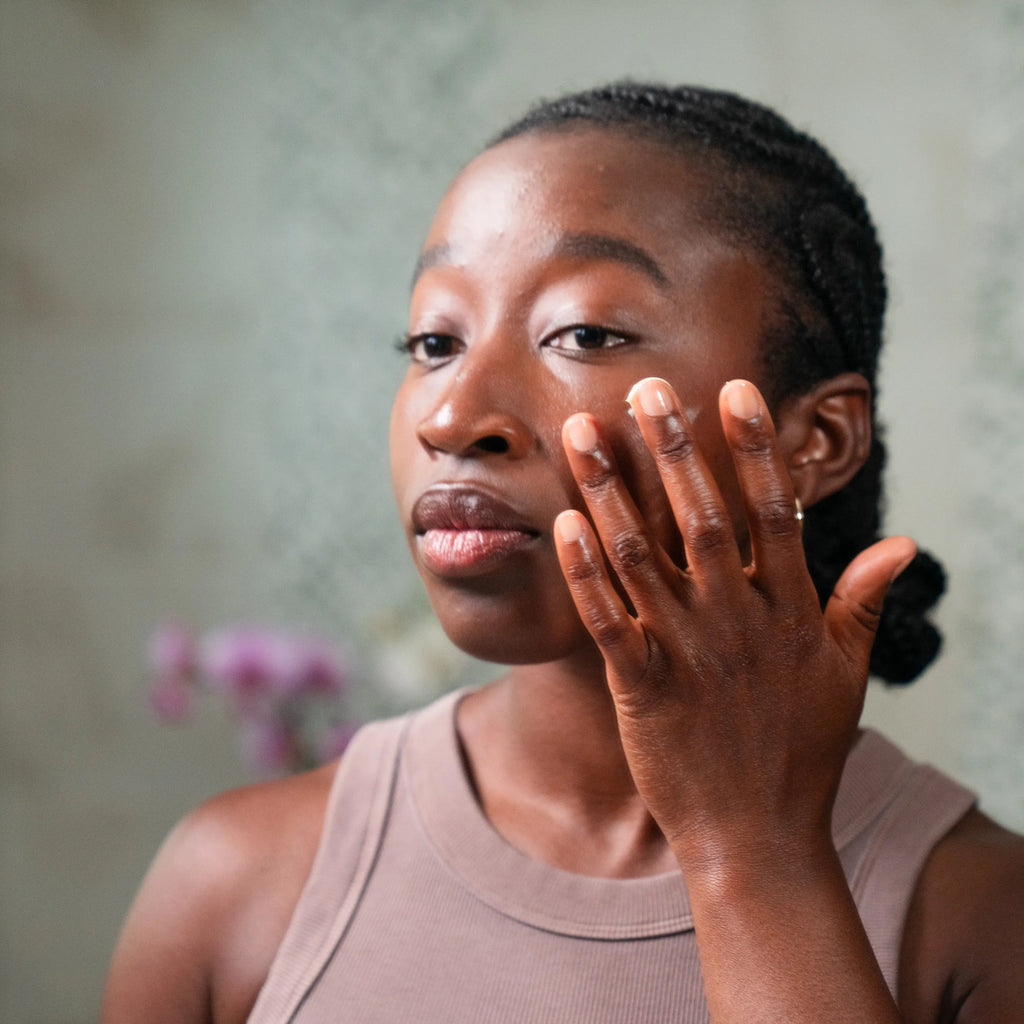 A woman wearing a taupe tank top is shown in a mirror applying cream on her face with her hand against a soft-focus indoor background.